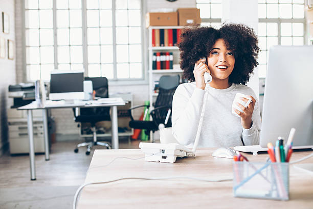 Smiling young businesswoman talking on the phone at workplace Cheerful young woman sitting on desk and talking on landine phone, holding a cup, looking away. Tools and mouse on desk. As background tall windows, desk with computer and seat, shelves with folders and boxes.. landline phone stock pictures, royalty-free photos & images