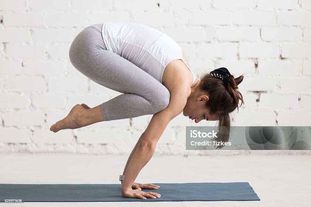 Yoga Indoors: Bakasana Beautiful young woman working out indoors, doing yoga exercise on blue mat, handstand asana, exercise for arms and wrists strength, Crane (Crow) Pose, Bakasana with arms straight, full length Activity Stock Photo