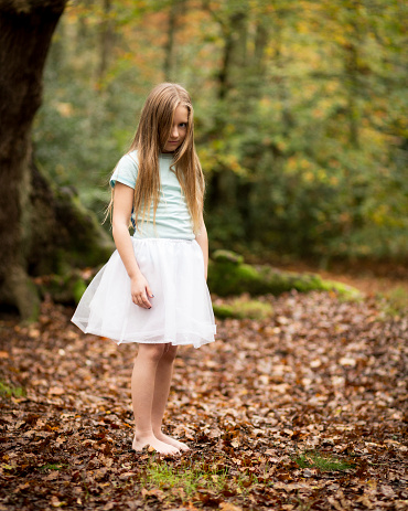 Portrait of a shy young child girl with long dark blond hair wearing a tutu skirt and bare legs and feet standing alone in a dark autumn forest clearing.