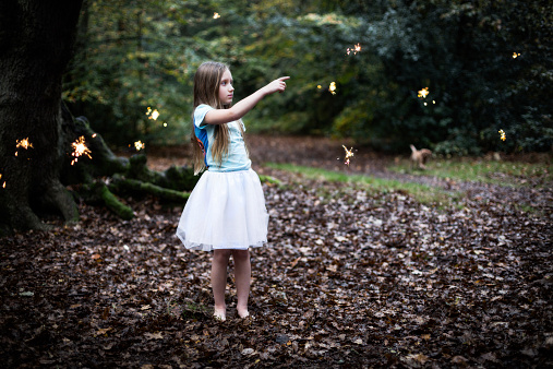 A young shy child girl with dark blond hair wearing a white tutu dress standing in a dark autumn forest surrounded by fairy sparkles pointing at one sparkle bare feet on a leave bed