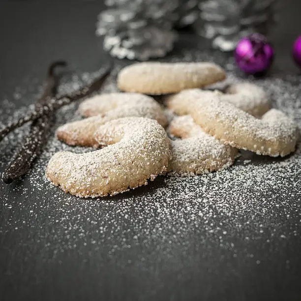 Freshly baked festive golden crescent cookies for Christmas and Advent sprinkled with icing sugar on a grey kitchen counter with Xmas decorations behind and shallow dof