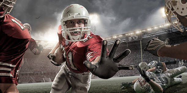 Close Up American Football Action Close up image of a professional American football player holding football and running with outstretched hand towards camera during American football game. Action is set in a generic outdoor floodlit American football stadium full of spectators under stormy evening sky. Players are wearing generic unbranded kit. Stadium advertising is fake.                                                                            defending sport stock pictures, royalty-free photos & images