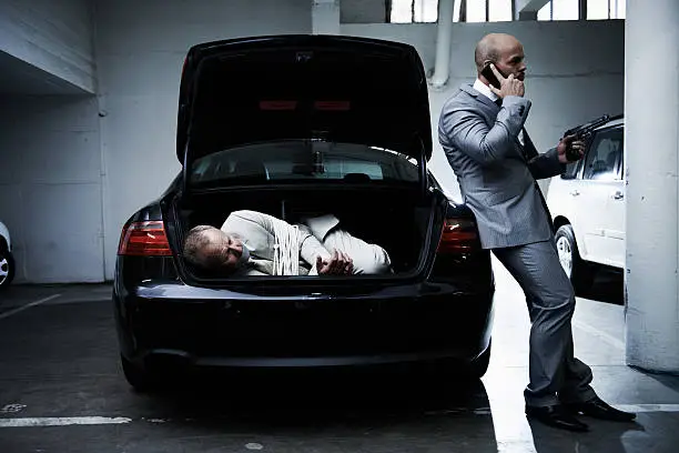 A frightened businessman lying in the trunk of a car while his armed captor talks on a cellphone