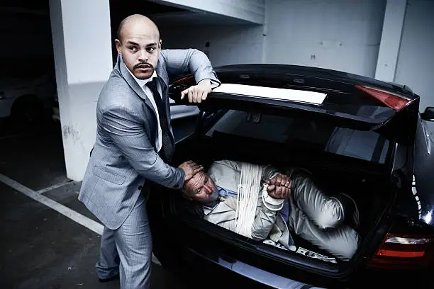 A gangster stuffing a bound and gagged businessman into the boot of his car