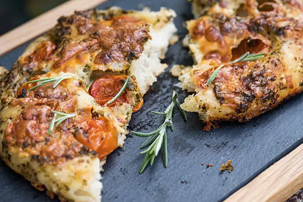 Vegetarian pizza.  Close up of a hand made focaccia bread topped with cherry tomatoes, pesto, cheese and herbs served on a slate serving platter.  Torn in half and garnished with fresh rosemary.