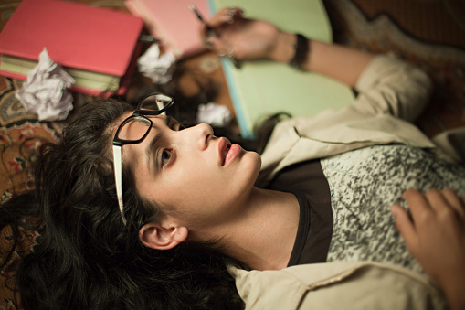 Indoor top view image shot in domestic room of a college student, late teen Asian girl reclining on floor carpet which is messed up with books, notebooks and paper balls. She is tired and relaxing while thinking something by looking away by putting eyeglass off and placed on head. One person, horizontal composition and selective focus.