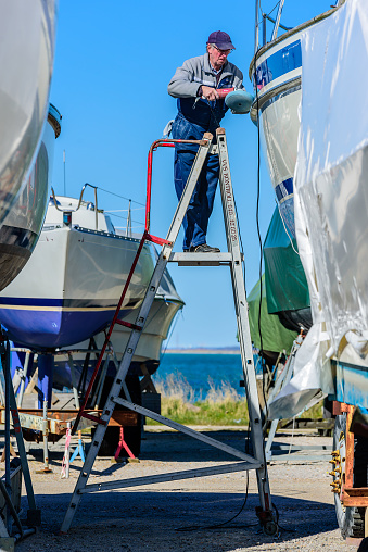 Falsterbo, Sweden - April 11, 2016: Senior adult man standing on a foldable ladder while polishing a sailboat. Real people in everyday life.