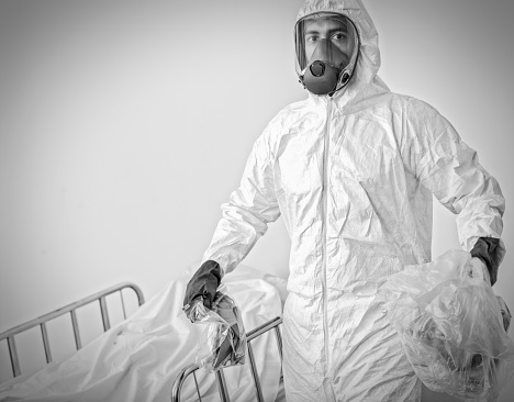 A man in a hazmat suit carries contaminated items away in plastic bags from a deceased patient in hospital.