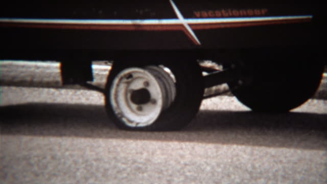 1971: Man fixing flat tire on camping trailer vacationeer vehicle.
