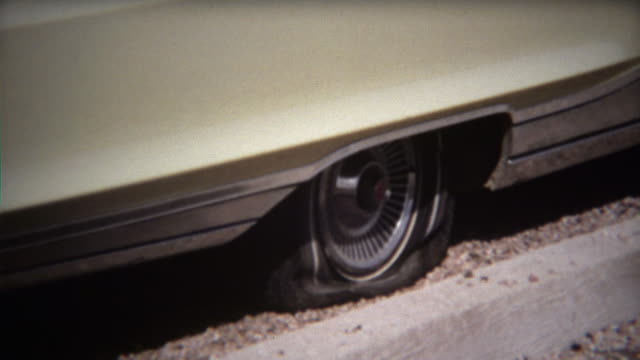 1971: Man fixes flat tire on white sedan car in hot sun.