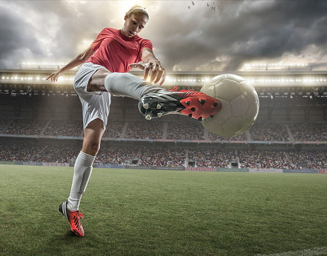 A close up image of woman soccer player kicking ball in a outdoor floodlit stadium full of spectators under a stormy evening sky. The player is wearing generic red and white unbranded football kit. The stadium is generic, created in Photoshop with fake advertising. Composite image with intentional lighting effects.