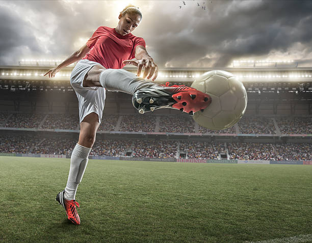 Girl Playing Soccer A close up image of woman soccer player kicking ball in a outdoor floodlit stadium full of spectators under a stormy evening sky. The player is wearing generic red and white unbranded football kit. The stadium is generic, created in Photoshop with fake advertising. Composite image with intentional lighting effects. kicking stock pictures, royalty-free photos & images