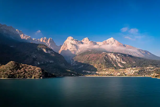 The Molveno lake with the alpine village of Molveno (TN) in long exposure under blue sky.
