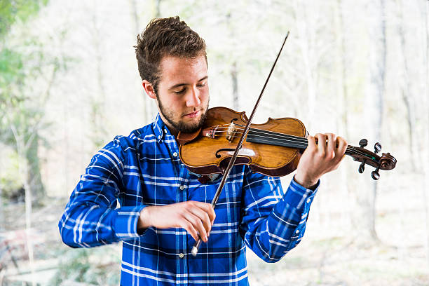 A violinist playing a violin or fiddle in front of a window over-looking a forest.