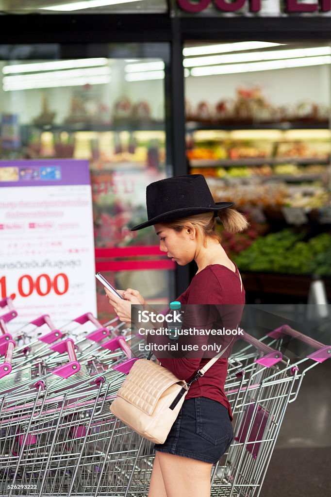 Thai girl with black hat and mobile Bangkok, Thailand - March 31, 2016: A thai girl with black hat and black hot pants is standing with mobile in front of a supermarket. Behind girl are pushcarts. In background is entrance to supermarket. Adult Stock Photo