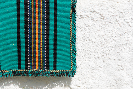 Woolen woven blanket in green and red color hanging over a white stucco wall in bright sunlight.