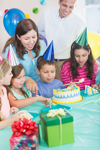 A little Hispanic boy having a birthday party with family and friends. He is turning 6 years old, sitting in the middle between his sisters, mother standing behind, reaching over to serve a slice of the cake. Dad and the rest of the children are watching.