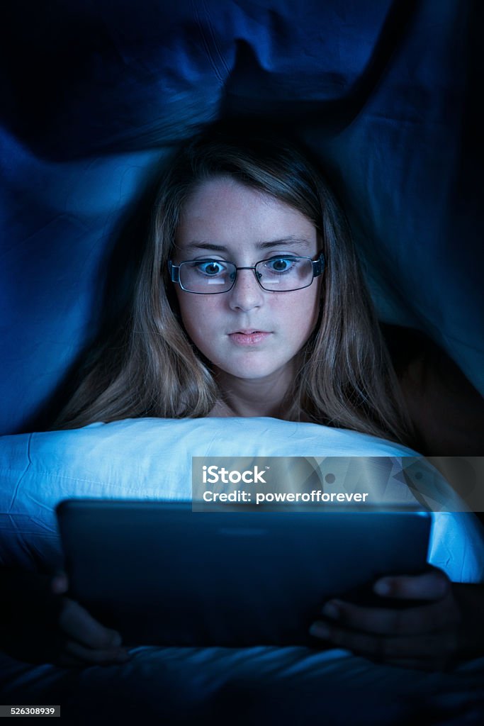 Teenage Girl using Tablet Computer under Bed Covers Teenage girl looking at tablet computer under the covers in her bed. Below Stock Photo