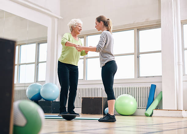 Trainer helping senior woman on bosu balance training platform Female trainer helping senior woman in a gym exercising with a bosu balance training platform. Elder woman being assisted by gym instructor while workout session. balance stock pictures, royalty-free photos & images