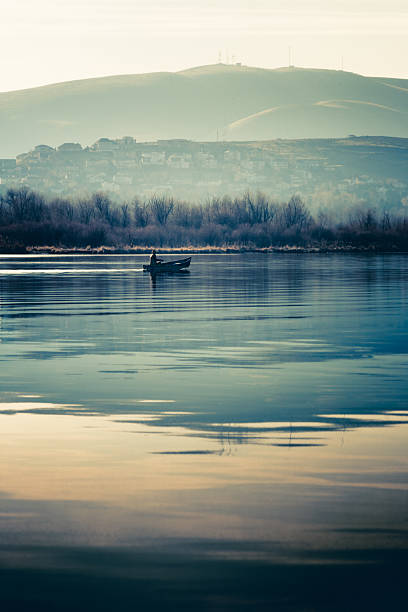A lone fisherman in a canoe gliding across tranquil waters of the Yakima Delta in Tri-Cities, Washington, where the Yakima River meets up with the Columbia River. Beautiful reflections ripple across the surface. Hills and residential community in the background. Soft, muted tones of blue and yellow. Color photograph with vertical composition and plenty of room for your copy at bottom of image.