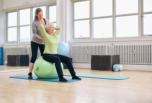 Senior woman sitting on a pilates ball  exercising at health club being assisted by her personal trainer. Physical therapist helping senior woman in her workout at gym.