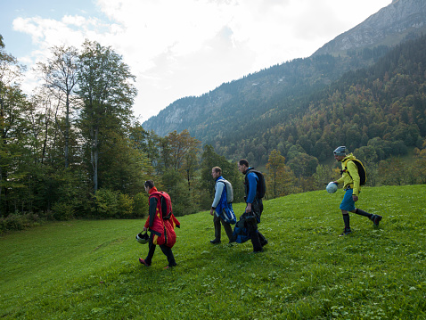 Wing suit fliers walk across meadow to jump, mountains & mist behind