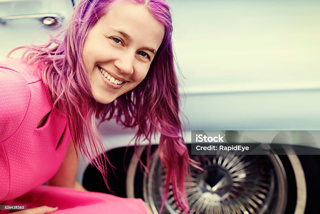 Cute purple-haired woman bends, smiling. by classic car A cute young woman with bright magenta hair and wearing pink smiles sweetly as she crouches next to  a car. Copy space on car hood. 20-29 Years Stock Photo