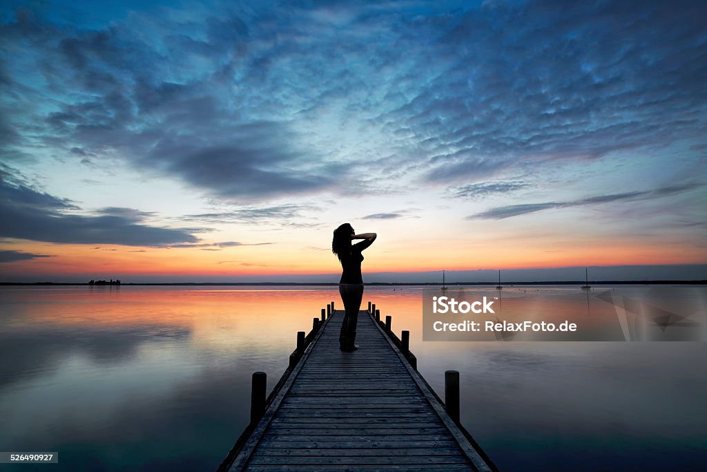 Silhouette of Woman on Lakeside Jetty looking at Sunset Cloudscape Young woman standing lakeside on jetty having a look at magical cloudscape and sunset colors.  Adult Stock Photo