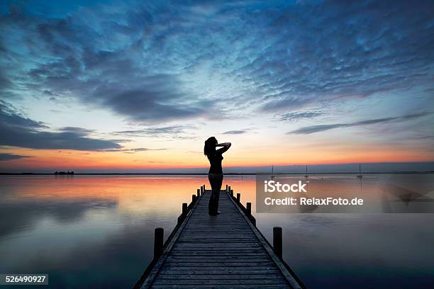 Silhouette Of Woman On Lakeside Jetty Looking At Sunset Cloudscape Stock Photo - Download Image Now