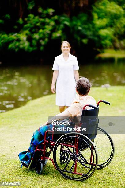 Smiling Nurse Approaches Woman In Wheelchair During Trip To Park Stock Photo - Download Image Now