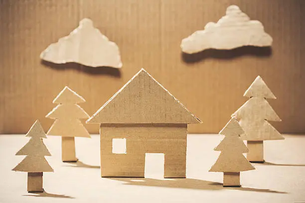 A simple diorama of a environmentally friendly recycling concept, depicting a house in a natural setting, surrounded by trees made entirely of brown cardboard paper.  Horizontal image.