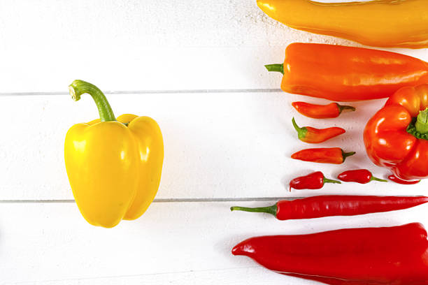 The image showcases a still life arrangement of various peppers on a textured, warm-toned surface. The composition includes a mix of red and orange bell peppers, along with a couple of long, slender red chili peppers. The peppers are grouped together, creating a visually appealing cluster with some overlapping. The lighting is dramatic, casting shadows that add depth and emphasize the textures of the peppers and the surface they rest on. The color palette is rich and warm, dominated by the vibrant reds and oranges of the peppers, which contrast with the more muted tones of the background. The overall impression is a classic still life, highlighting the natural beauty of the produce.