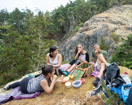 Teenaged girls and father taking a break from backpacking and enjoying healthy vegetarian lunch of fruit, berries, vegetables, hummus, crackers and juice.  Multi-ethnic family (Asian and Caucasian) with forest, cliffs and mountain in the background.  Wilderness park near Victoria, Vancouver Island, British Columbia, Canada.
