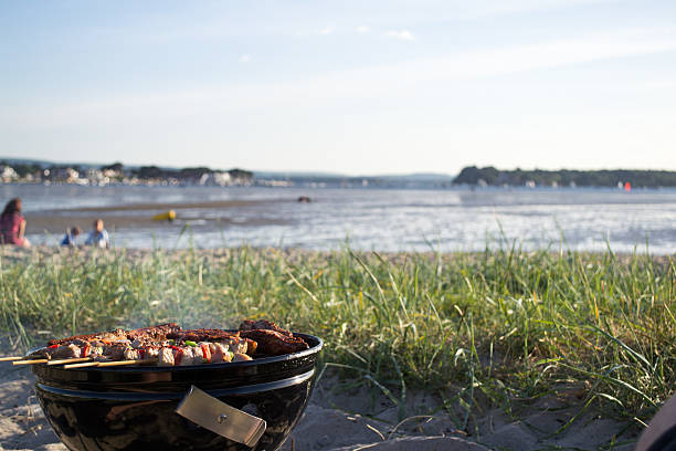 BBQ food cooking on the beach with family in background A BBQ with kebabs cooking on a sandy dune in summer. A family is in the background with the BBQ and food in the foreground. Grass covers part of the dune and the harbor is at low tide. family bbq beach stock pictures, royalty-free photos & images