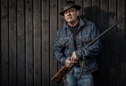 Man in jeans jacket and hat posing with a shotgun in the afternoon sun infront of a wooden wall.