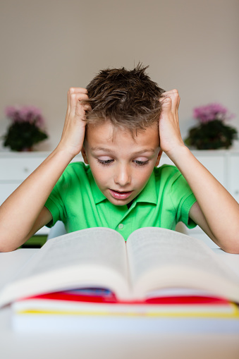 Young boy in green polo shirt having serious learning difficulties while trying to read a textbook from school.