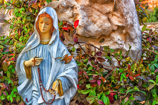 Statue of the Blessed Virgin Mary with wooden prayer beads necklace in a house rock  garden  with treesin  Medjugorje