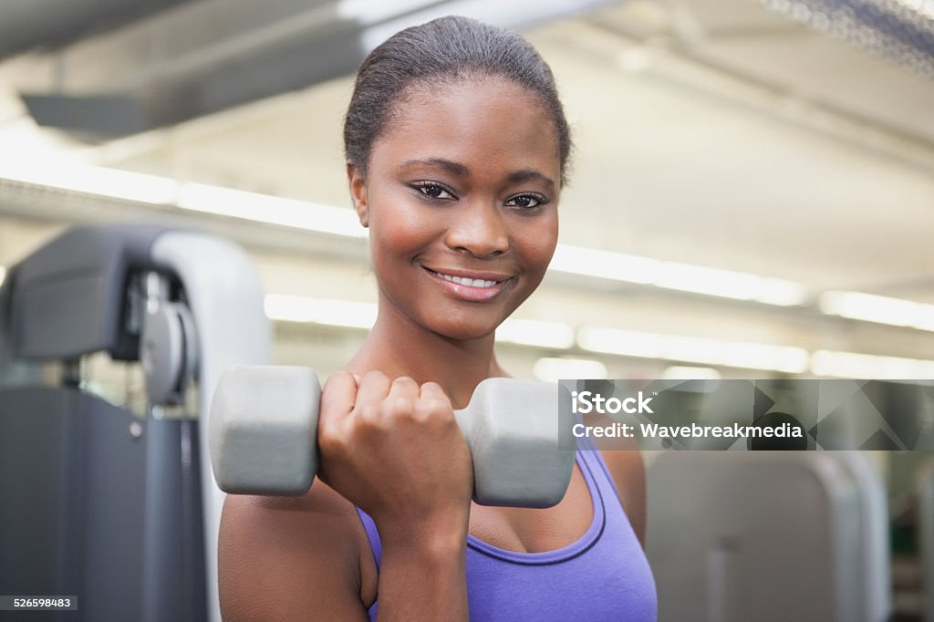 Fit woman smiling at camera holding dumbbell Fit woman smiling at camera holding dumbbell at the gym Dumbbell Stock Photo