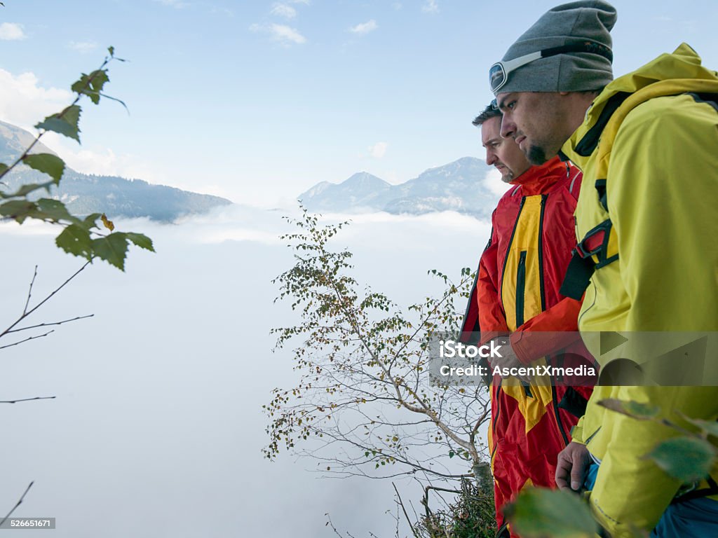 BASE/Wing suit fliers ponder landing before jumping BASE/Wing suit fliers ponder landing before jumping from cliff, mountains & fog behind Above Stock Photo