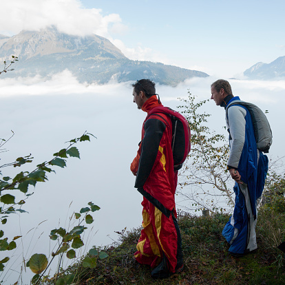Wing suit fliers ponder landing before jumping from cliff, mountains & fog behind