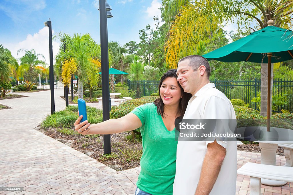 Couple take a selfie in the park A couple is taking a selfie with a cell phone.  They are in a park.   They are enjoying a beautiful summer day.  They are in their thirties. She has long black hair and he has a buzz cut.  Shot with Canon 5D Mark 3.  rr Adult Stock Photo