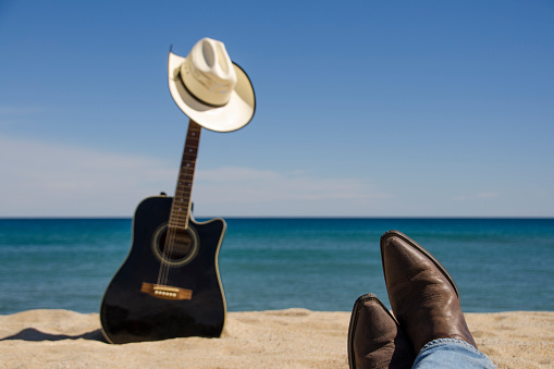 A non focused black acoustic guitar and a white cowboy hat on the beach