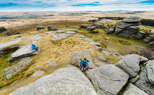 A lady climbs up a rocky outctrop (Bellever Tor) on Dartmoor, Devon, UK. A man sits on a rock in the background watching.