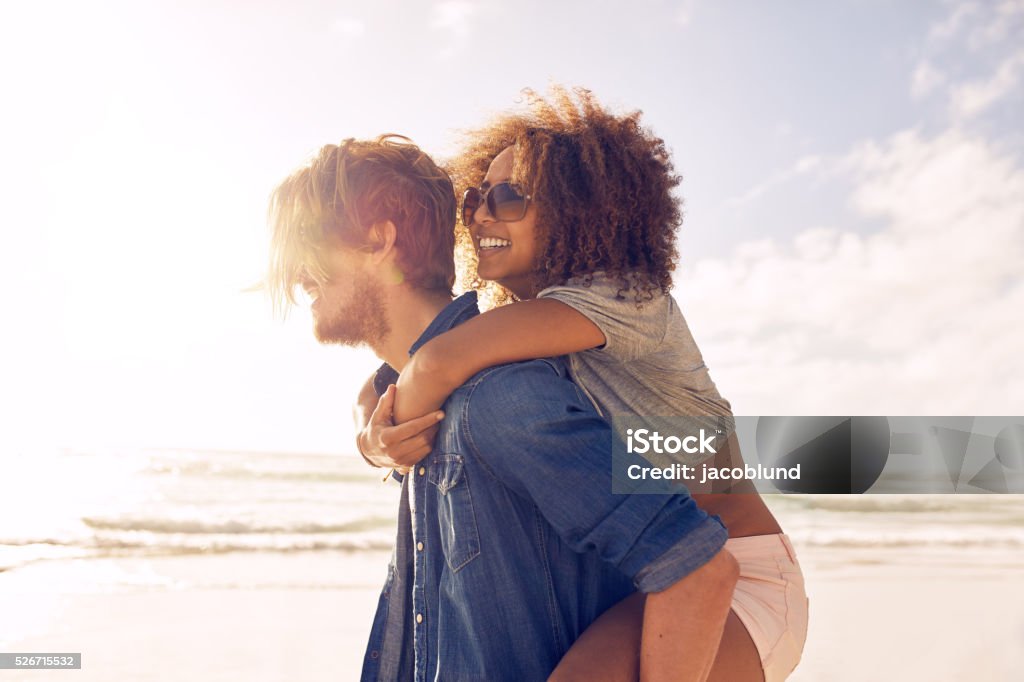 Young couple enjoying their summer vacation on beach Side view portrait of young man carrying his girlfriend on his back at the beach. Boyfriends giving piggyback ride to his beautiful girlfriend at seashore. Couple - Relationship Stock Photo