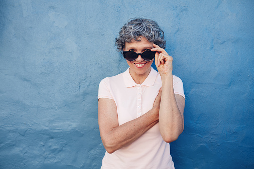 Portrait of smiling mature woman peeking over sunglasses against blue background. Beautiful middle aged female looking at camera.