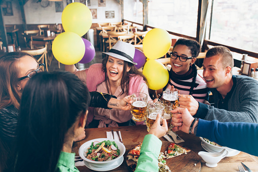 Celebratory toast with friends in restaurant. Young girls and boys drinking beer and eating pizza and salad together. One girl with eyeglasses and braids, other with white hat. Tall windows, balloons and wall with pictures as background.