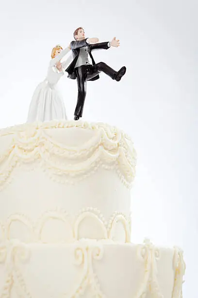 Humorous wedding cake toppers, with the bride chasing and catching the terrified groom who is escaping, running from getting married. A fun bridal shower, renewed vows, or destination wedding reception joke for newlyweds or marriage anniversary couples. Frosted dessert with husband and wife figurines on a white background, with copy space.