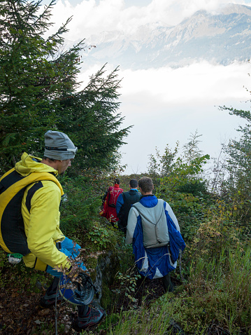 Wing suit fliers walk through forest to launch site, mountains & mist behind