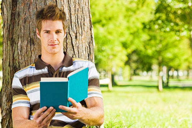 istock Smart young male reading book sitting by tree 526746391