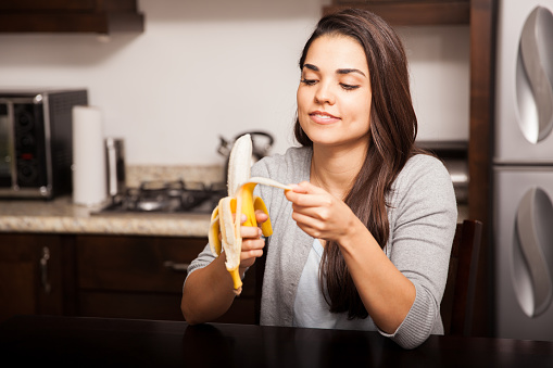 Pretty young brunette peeling a banana before eating it while sitting in the kitchen